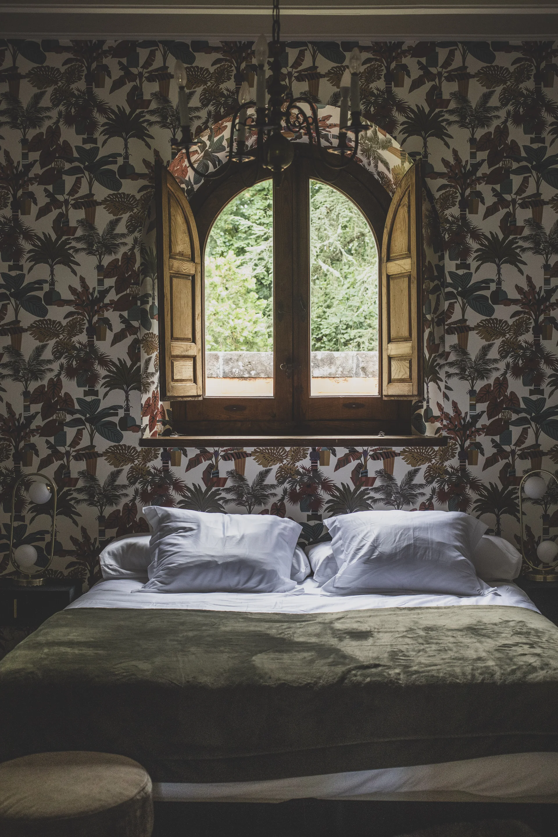 Cama bajo una ventana en arco, rodeada de un papel mural de vegetación.